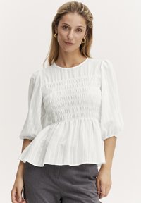 Vit blus med smockad byst, strukturerade ränder, puffärmar och peplumslut. Buren med grå byxor, med rund halsringning.
