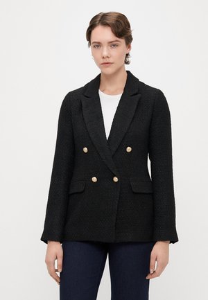 Sort tweed dobbeltradet blazer med guldbeslag, klapkrave, to forlommer og tekstureret stof. Bæres over en hvid skjorte.