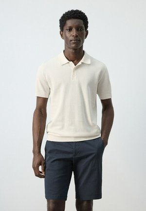 Hombre con una camisa tipo polo de manga corta color crema y pantalones cortos azul marino, de pie contra un fondo claro y liso.