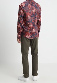 Chemise à manches longues à fleurs multicolores avec des nuances de rouge, marine et vert, associée à un pantalon vert olive et des baskets blanches, vue de dos.