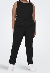 Kvinna iklädd svart ärmlös topp, svarta byxor med dragsko i midjan och vita sneakers som står mot en enfärgad ljus bakgrund.