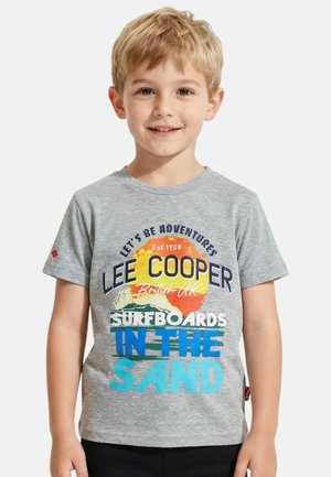 Jeune garçon blond portant un t-shirt gris avec un texte coloré sur les planches de surf et les aventures, souriant et posant devant un fond blanc.