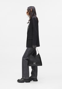 Woman in black blazer and gray jeans holding a black triangular handbag with silver "K" logo, standing sideways on a white background.