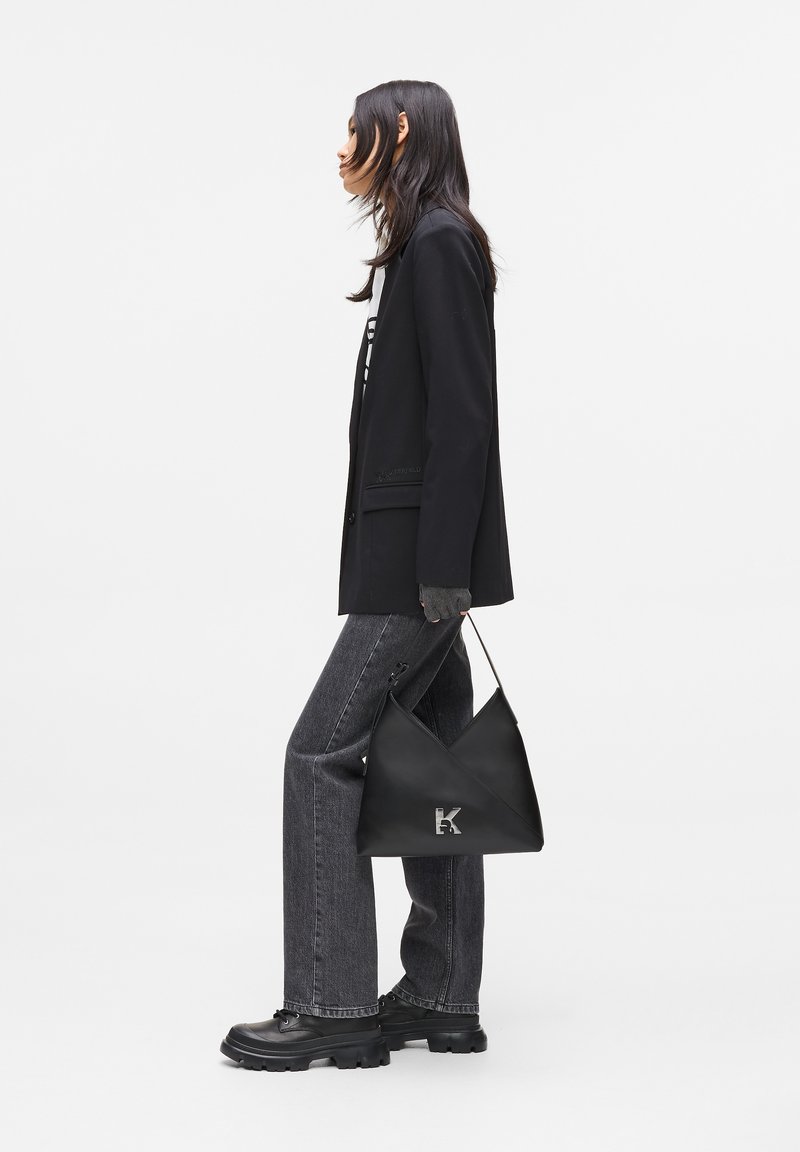 Woman in black blazer and gray jeans holding a black triangular handbag with silver "K" logo, standing sideways on a white background.