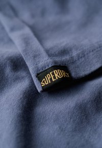 Navyblåt stof tæt på med en tekstureret overflade, med et sort mærke, der viser "SUPERDRY" i gyldne bogstaver syet i kanten.