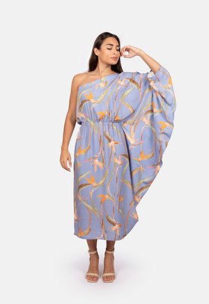 Modèle portant une robe fluide à une épaule avec un imprimé floral bleu, se tenant avec assurance, une main sur la hanche et une expression légère.
