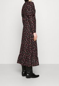 Kvinna i en svart lång blommig klänning med puffärmar och volangkant, i kombination med svarta cowboystövlar, stående åt sidan.