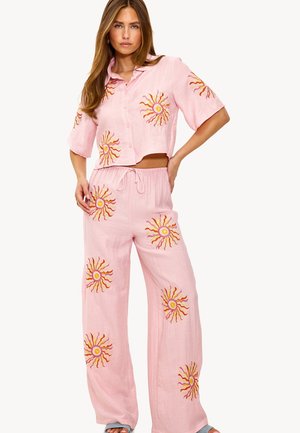 Chemisier rose coupé court avec broderie de soleil, associé à un pantalon large présentant des motifs similaires. Tissu léger, coupe décontractée et taille à cordon.