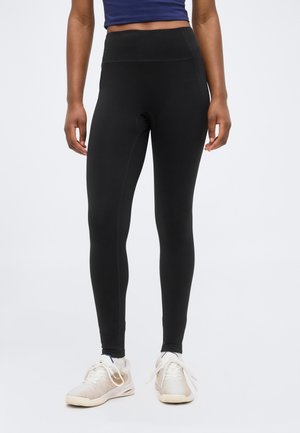 Osoba oblečená v čiernych legínach s vysokým pásom a bielych športových topánkach, stojaca pred jednoliatym svetlosivým pozadím.