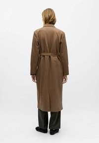 Una mujer con cabello rubio lleva un abrigo largo marrón con cinturón, pantalones oscuros de corte ancho y zapatos negros, de pie frente a un fondo blanco.