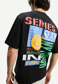 Black cotton t-shirt featuring colorful graphic prints, including text and a yellow tennis ball design. Relaxed fit with short sleeves.