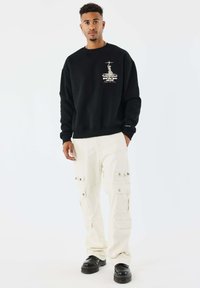 Man in zwarte sweatshirt met grafisch ontwerp, crème cargo-broeken en zwarte platform schoenen, staande tegen een witte achtergrond.