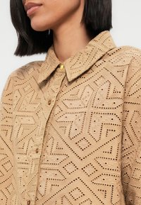 Chemise beige avec des motifs découpés complexes, dotée d'un col, d'un devant à boutons, et de petits boutons dorés en accent. Tissu texturé.