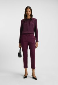 Langarmbluse und schmal geschnittene Hose in tiefem Lila. Die Bluse hat einen gerafften Kragen; die Hose hat eine glatte Textur. Schwarze Slingback-Absätze und eine Handtasche.