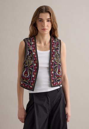 Junge Frau trägt eine ärmellose schwarze Weste mit bunter Blumenstickerei über einem weißen Tanktop und schwarzen Hosen, steht vor einem einfarbigen Hintergrund.