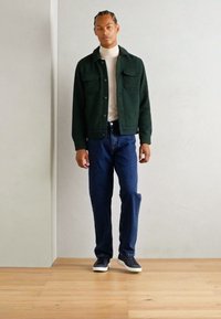 Dark green textured jacket with two front pockets, white turtleneck, blue jeans, and black sneakers with white soles. Standing on wooden floor.
