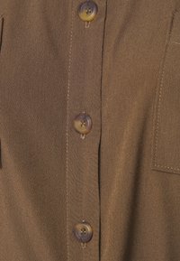 Chemise marron à boutons avec une texture lisse, dotée de boutons translucides et d'une poche poitrine unique sur le côté gauche.