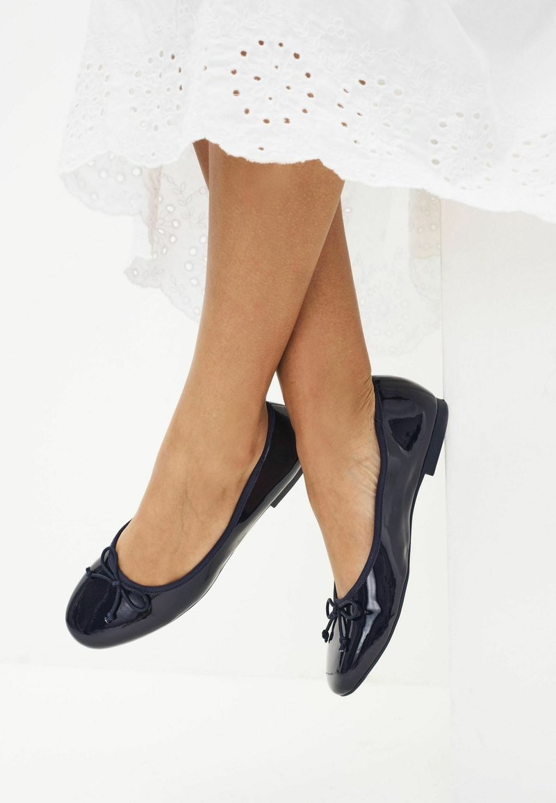 Pieds chaussés de ballerines noires brillantes avec de petits nœuds, croisés aux chevilles, sous une robe blanche en broderie anglaise.