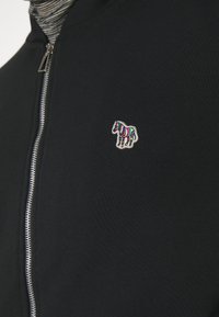 Veste noire à fermeture éclair avec un écusson multicolore en forme de cheval rayé sur la poitrine, portée par-dessus une chemise rayée grise et noire.