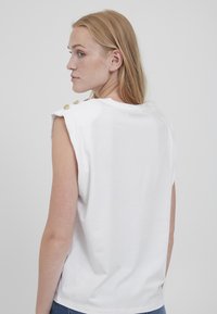 Hvid bomuld t-shirt med korte ærmer, der har guldknapdetaljer på skulderen. Glat tekstur, enkelt design. Vist fra bagenden.