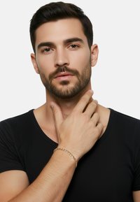 Gouden armband om een hand met een zwart shirt met korte mouwen. De armband heeft ronde schakels die in een eenvoudig patroon zijn gerangschikt.