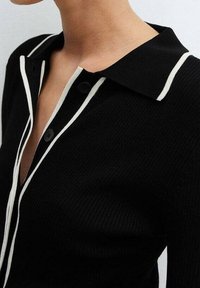 Camisa negra de canalé con un cuello blanco ribeteado y cierre de botones. La tela parece texturizada con un diseño ajustado.