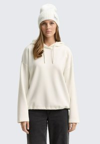 Joven con cabello largo que lleva un gorro blanco, sudadera blanca holgada y pantalones negros, de pie contra un fondo gris claro liso.