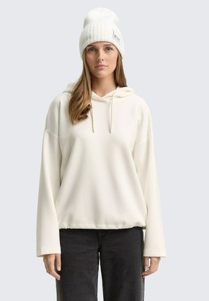 Ung kvinna med långt hår som bär en vit mössa, en lös vit hoodie och svarta byxor, stående mot en enkel ljusgrå bakgrund.