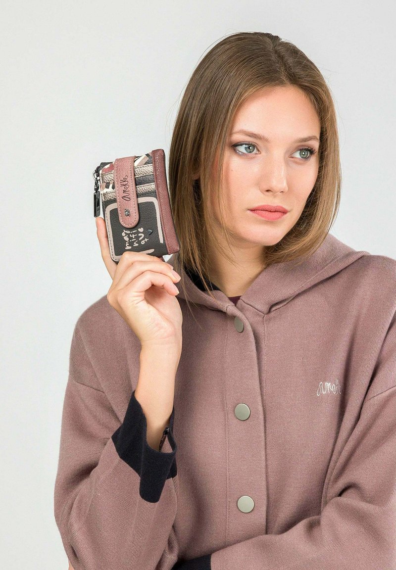 Cartera texturizada en marrón oscuro y claro con una correa rosa, que presenta un cierre de cremallera y múltiples espacios para tarjetas. El modelo la sostiene contra un fondo gris.