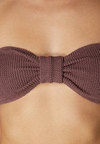 Brun vävd bandeau-bikinitop med en texturerad yta och en central rosett. Slät hudton bakgrund som framhäver produktens design.