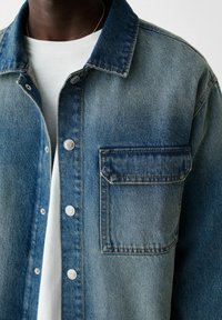 Camicia in denim di colore azzurro chiaro con davanti a bottoni, colletto a punta e una tasca sul petto. Presenta un tessuto testurizzato e cuciture visibili.