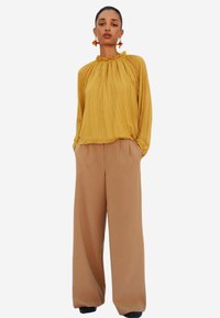 Blusa gialla a maniche lunghe con pieghe e scollatura arricciata, abbinata a pantaloni a gamba larga beige a vita alta. Design minimalista, texture morbida.