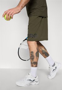 Iš dalies matomas asmuo, laikantis teniso kamuoliuką ir raketę, vilkintis alyvuogių spalvos šortus, baltus sportinius batus ir kojines, turintis tatuiruotes ant abiejų blauzdų.