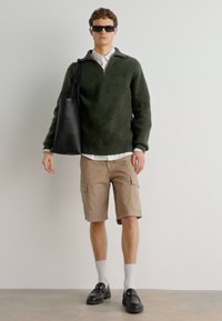 Pullover verde scuro a coste con mezza zip, pantaloni cargo beige, borsa a tracolla nera, occhiali da sole rotondi e scarpe nere con hardware decorativo.
