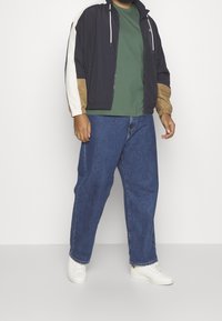 Marinduk, beige och kremfärgad färgblockerad jacka med dragsnören; grön t-shirt; blå avspänd jeans; vita sneakers.