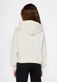 Persona con lunghi capelli castani che indossa una felpa bianca semplice e pantaloni neri, vista da dietro su uno sfondo bianco.