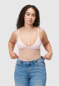Jasnoróżowy biustonosz typu bralette z cienkimi ramiączkami w połączeniu z beżową bielizną modelującą. Modelka nosi luźne niebieskie jeansy o średnim stanie, prezentujące swobodny krój.