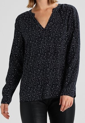 Kobieta w czarnej, długorękawowej bluzce z białym, nieregularnym wzorem kropek i delikatnym dekoltem w serek, w połączeniu z czarnymi spodniami.