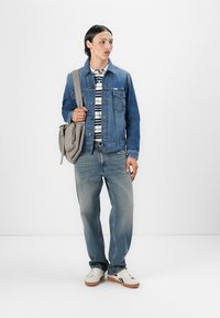 Denimjakke med frontlommer, stripete skjorte, lyseblå jeans, beige veske og hvite sneakers med brune såler, satt mot en ensfarget bakgrunn.