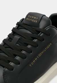 Pantof sport din piele neagră cu șireturi negre, având logo auriu pe limbă și pe lateral. Vârf rotunjit și talpă albă contrastantă.