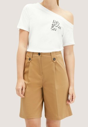 Kobieta ubrana w białą, odkrytą na ramionach koszulkę z napisem "życie toczy się dalej" oraz wysokie, beżowe szorty z guzikami, stojąca na neutralnym tle.