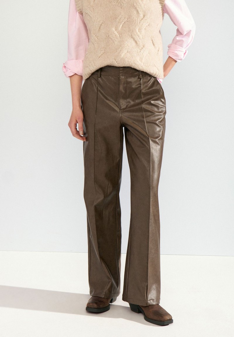 Personne portant un pantalon flare en cuir marron, des bottes marron foncé, un gilet en tricot beige et des manches de chemise roses retroussées, debout devant un fond clair.