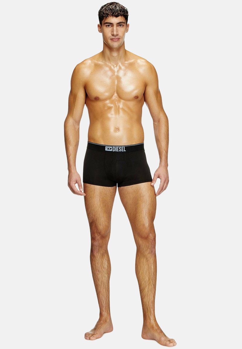 Μαύρο βαμβακερό μποξεράκι με μπλε λογότυπο στη ζώνη, εφαρμοστό σχέδιο και μήκος κάτω από το γόνατο, που μοντελοποιείται από ένα γυμνό άνδρα.