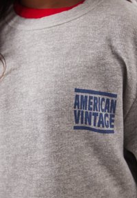Personne portant un sweat-shirt gris avec un col rouge et le texte bleu « AMERICAN VINTAGE » imprimé sur la zone de la poche poitrine.