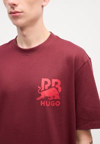 Maroon cotton t-shirt featuring a large red graphic logo depicting a bull and text "RB HUGO" on the left chest area. Round neckline.
