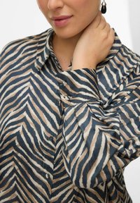 Koszula z guzikami uszyta z lekkiego materiału, z zygzakowatym wzorem zebry w ciemnym niebieskim, beżowym i białym kolorze, z kołnierzykiem i mankietami.