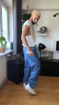 Mlad moški z blond lasmi in brado, oblečen v brezrokavnik bele barve in ohlapne modre kavbojke, stoji na prstih v svetli, moderni sobi.