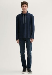 Marineblauer Pullover mit Stehkragen, langen Ärmeln und Kordelzugdetails, kombiniert mit dunkelblauen Jeans und schwarzen Sneakers.