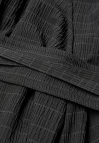 Tissu noir avec un motif en quadrillage texturé et une ceinture assortie posée dessus, mettant en valeur les plis doux et les détails de couture du matériau.