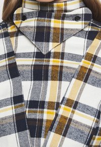 Plaid shirt featuring navy, white, and yellow checks. High collar with a pointed design and button accents. Soft fabric texture.